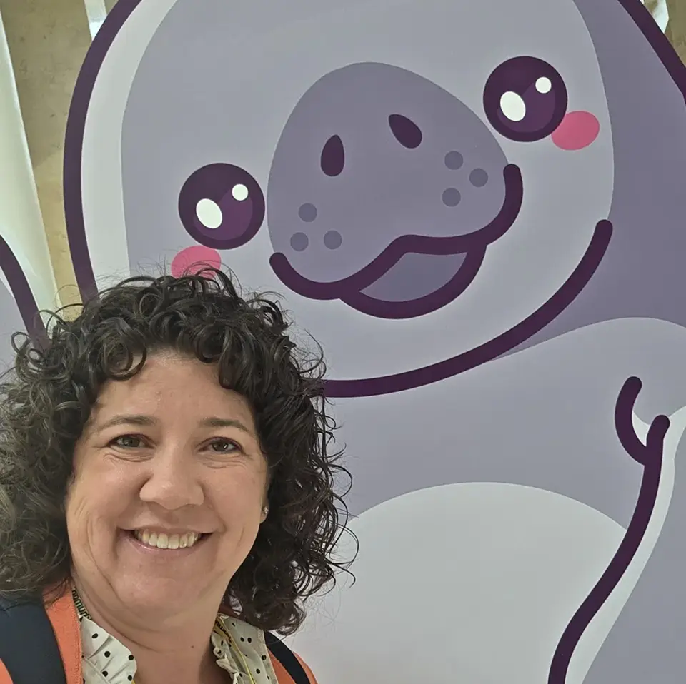 A person with curly hair is smiling in front of a large, cartoon-style illustration of a happy animal, likely a seal or sea lion, with a friendly expression. The background features light colors, enhancing the cheerful scene.