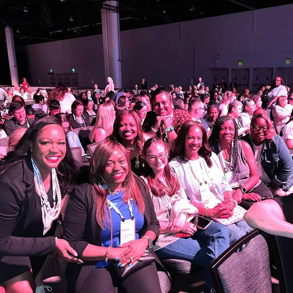 A group of eight women is smiling and posing for a photo while seated at a conference event. The women are dressed in various outfits, with some wearing necklaces and badges. In the background, there are many attendees seated in rows, indicating a lively atmosphere. One woman in front prominently displays a name tag that reads "Anitra." The lighting gives a pinkish hue to the scene, enhancing the cheerful environment.