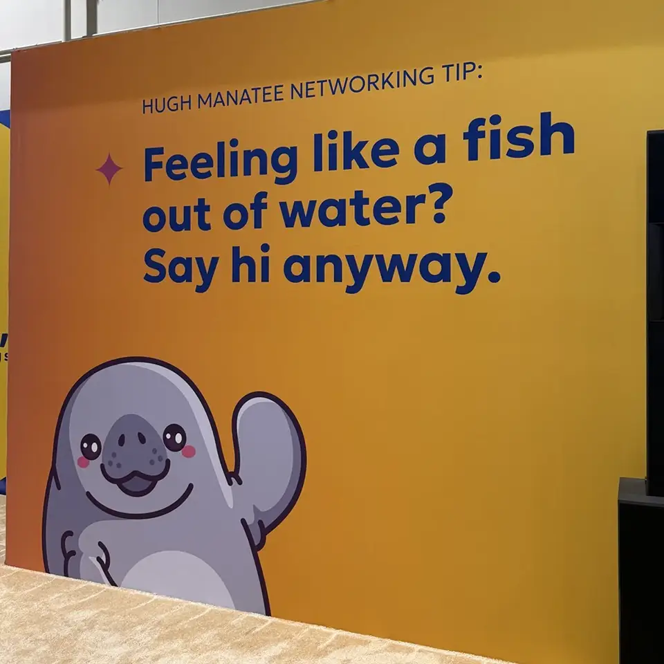 A playful wall display featuring a cartoon manatee along with a networking tip. The background is bright yellow, and the text reads, "HUGH MANATEE NETWORKING TIP: Feeling like a fish out of water? Say hi anyway." The manatee has a friendly smile and a raised flipper, adding a whimsical touch to the advice.