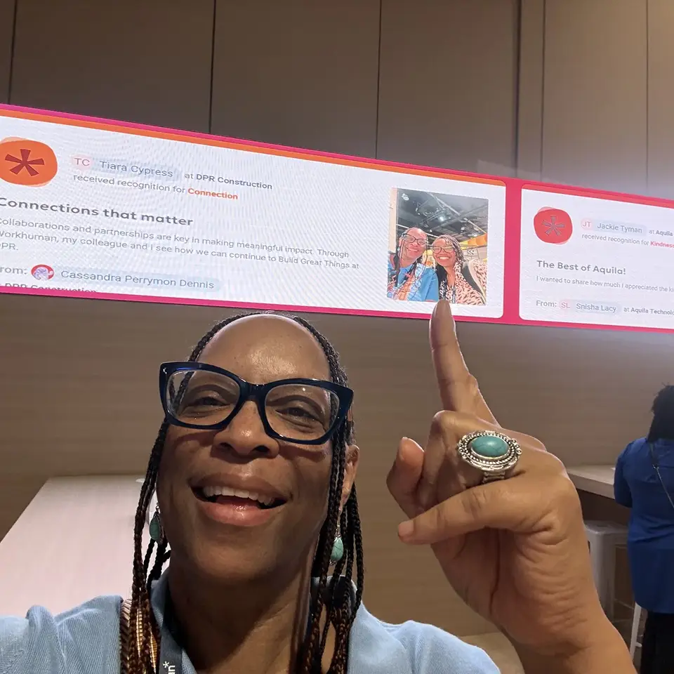 A woman with braided hair and glasses smiles and points at a digital screen displaying recognition messages for her colleagues. The screen shows that TC Tiara Cypress at DPR Construction received recognition for "Connection," and JT Jackie Tyman at Aquila was recognized for "Kindness." Below, a message reads, "Connections that matter," emphasizing the importance of collaboration and partnerships in making a meaningful impact.