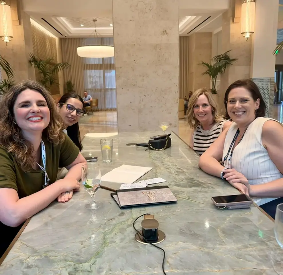 A group of four women smiles while seated at a marble table in a bright, modern venue. They are enjoying drinks, with one glass containing a lime wedge. In the background, plants and soft seating create a welcoming atmosphere, and a few people can be seen in the distance. The scene conveys a relaxed and enjoyable gathering.