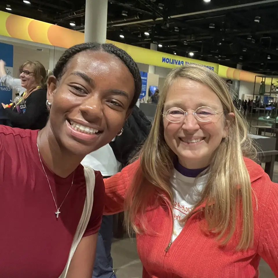 A young woman and an older woman are smiling together in a selfie. They are indoors, with a spacious exhibition area visible in the background. The young woman wears a red shirt and a necklace, while the older woman has long hair and glasses, dressed in a red jacket over a white shirt. People can be seen milling around in the background.