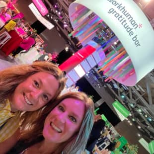 Two women smile for a selfie in front of a colorful display featuring the "Workhuman Gratitude Bar." The scene is vibrant, with pink and green decorations in the background and hanging ribbons from a prominent overhead sign.
