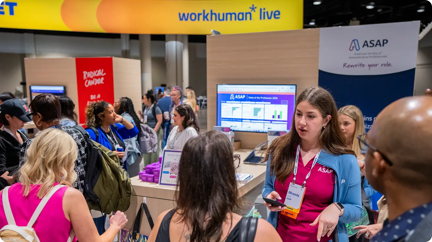 A woman wearing a pink shirt and a badge labeled "Miranda" stands at a booth displaying the logo "ASAP" while speaking to a group of people. In the background, a large sign reads "workhuman live," and a screen shows various charts under the title "State of the Profession 2023." The booth is surrounded by attendees engaged in conversation.