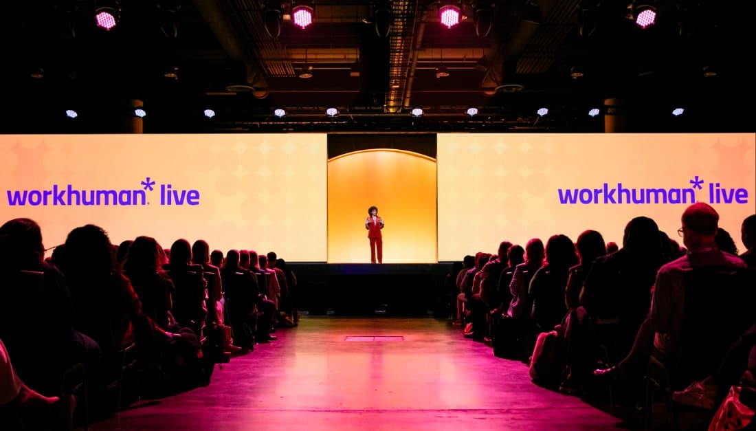 A speaker stands on a stage in front of large screens displaying the text "workhuman* live" against a bright orange background. An audience is seated in rows facing the stage, with some audience members visible in the foreground. The lighting is dim in the venue, highlighting the speaker and the screens.