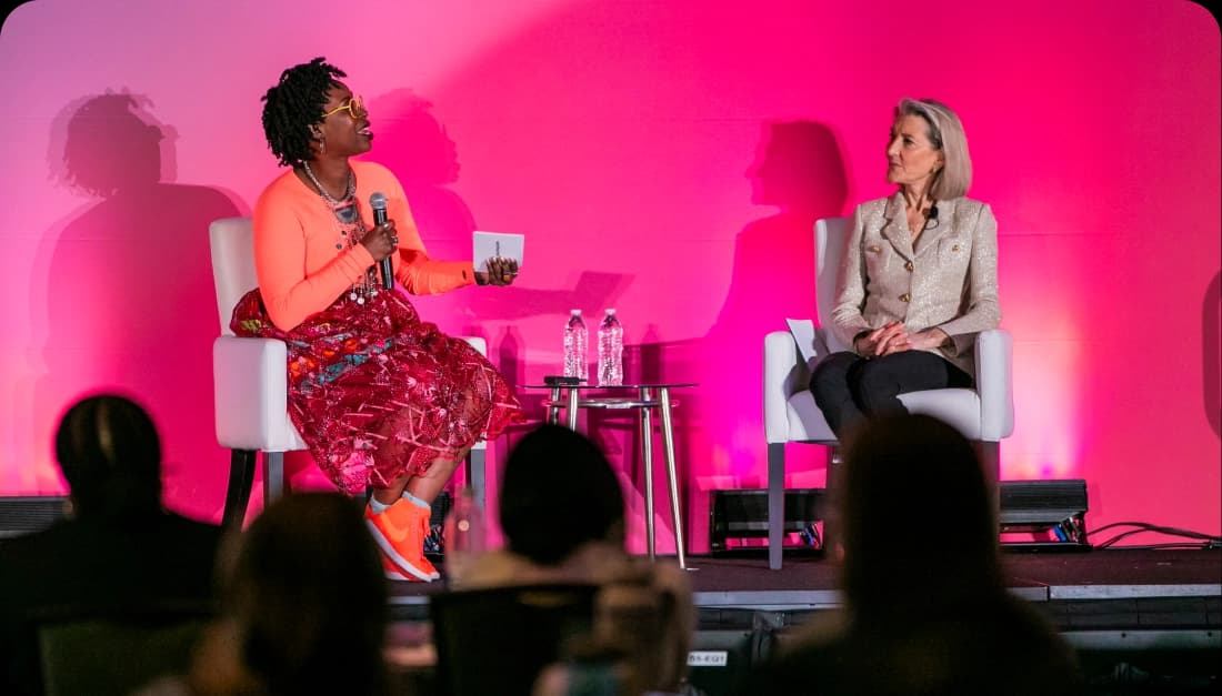 A person wearing a bright orange top and a colorful skirt speaks into a microphone while seated on stage next to another person in a light-colored blazer. They are both facing an audience, with a pink backdrop behind them. On a small table between them, there are two water bottles.