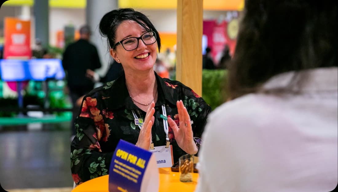 A person wearing glasses and a floral shirt is gesturing with their hands while smiling during a conversation at a table. A sign on the table reads "OPEN FOR ALL," and there are snacks visible in front of them. The background shows a vibrant, busy event space.